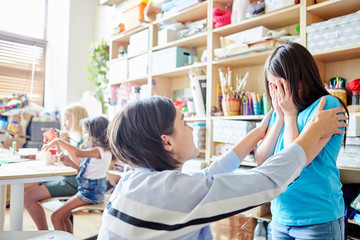 Side view of young female teacher comforting crying girl during lesson in art school