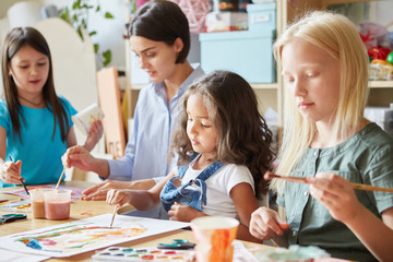 Fototapeta premium Young teacher and sweet schoolgirls sitting at desk in art school and painting bright pictures together