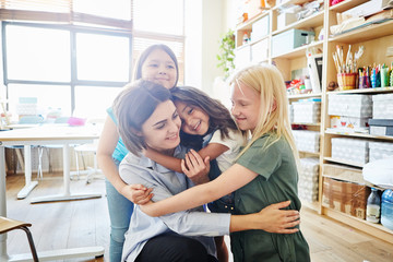 Cute girls smiling and embracing tenderly their young teacher in classroom at art school