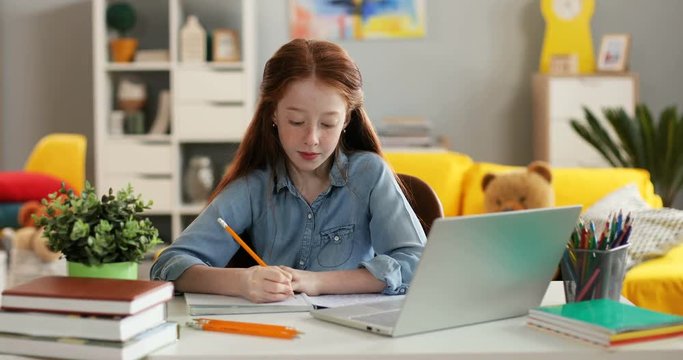 Portrait shot of the pretty Caucasian red haired teen girl writing exercise of homework in the copybook and thinking while sitting at the table with a laptop computer at home.