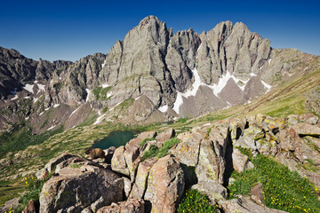 Marmot sits on rocks on a saddle of Humboldt Peak wtih Crestone Needle and Crestone Peak in distance rising above South Colony Lake, Colorado