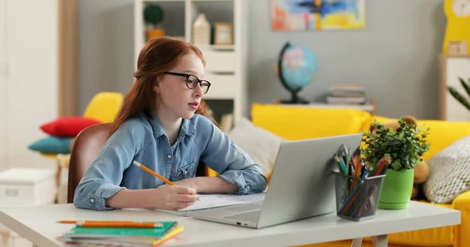 Caucasian teen cute red haired girl in glasses writing something while doing homework at the table with laptop computer in the cozy room.