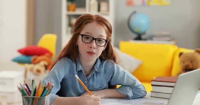 Portrait of the Caucasian teen cute red haired girl in glasses writing something while doing homework at the table with laptop computer and then smiling to the camera in the cozy room.