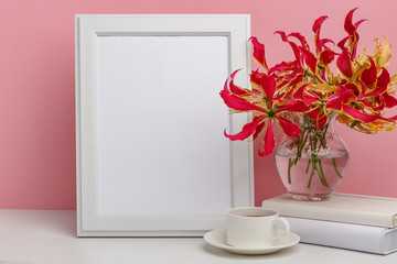 Composition from a decorative wooden frame and red and yellow flowers of Glorios in a glass vase on a white shelf on a pink background