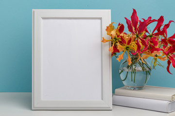 Composition from a decorative wooden frame and red and yellow flowers of Glorios in a glass vase on a white shelf on a blue background