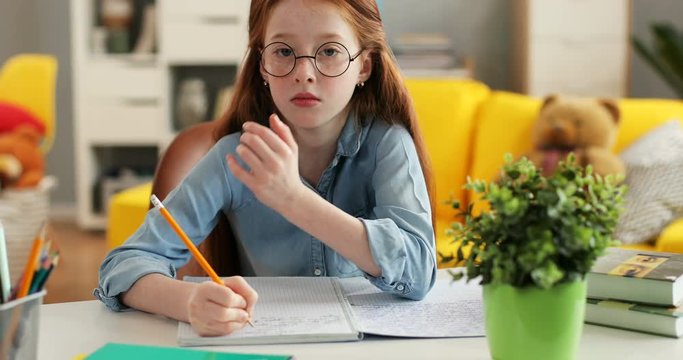 Close up of the nice teenage girl with red hair and in glasses writing exercise in the copybook as doing her homework in the nice room, then smiling to the camera. Portrait.