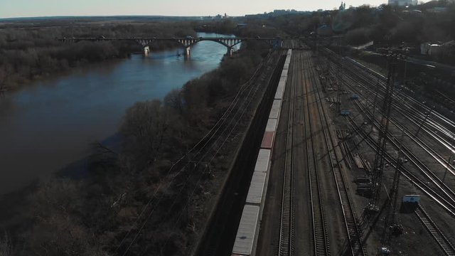 Aerial view of a large train on the rails next to the river. The camera flies over a long train of tanks standing on the rails
