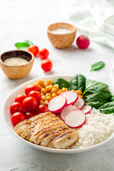 Healthy buddha bowl with spinach, tomato, chickpea, rice, radish and grilled chicken on concrete background. Selective focus.
