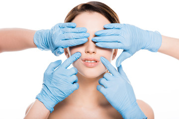 cropped view of plastic surgeons in blue latex gloves covering eyes and touching face of girl isolated on white