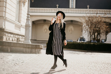 Young attractive lady with blonde straight hair, sunglasses, coat and hat is walking in the city with coffee to go