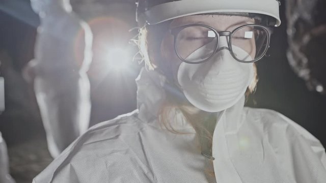 Portrait of a female biologist in a protective mask and a white dressing gown in glasses, which, with rude captives, explores an underground facility.