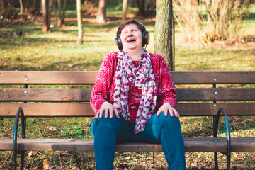 Laughing elder lady sitting on a bench in the park listening to music on her big black headphones – Excited and happy old lady outdoor on a bright sunny day