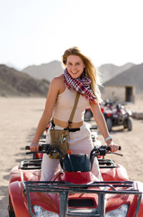 Fototapeta na wymiar Young attractive woman with long blonde hair is sitting on the quad bike, windy sunny day. Travelling, extreme mood, desert on the background
