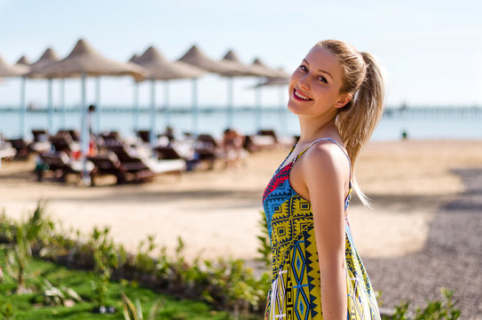 Young woman with ponytail, blonde hair is looking on the sea horizon, vacation mood, summer travel concept. Beach umbrellas on the background
