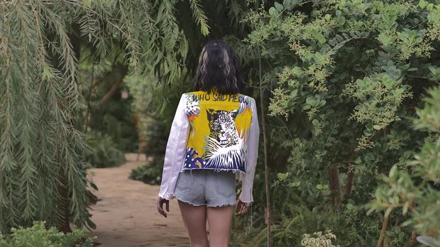 Slender girl in short shorts and a white jacket with a picture of a leopard on the back walks in the botanical garden, the tropics. She looks at the camera while turning