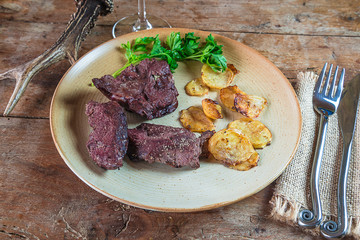 Deer meat roasted with potatoes on the rustic plate, cutlery and deer antler on the old wooden table