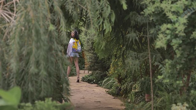 Slender girl in short shorts and a white jacket with a picture of a leopard on the back walks in the botanical garden, the tropics. She looks at the camera while turning