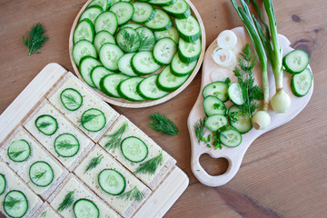 Lekkie, wiosenne kanapki z serem, świeżym ogórkiem i koperkiem, obok taca z plasterkami ogórka i deska pełna zielonych wiosennych warzyw