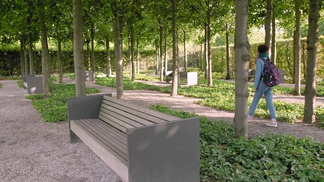 Hannover, Germany. A young female tourist with a map in her hands and a backpack walks along the alleys of the park at sunset time. In the frame a lot of trees and stone benches. Goes back to the