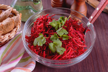 Thinly sliced ​​fresh beets in a glass salad bowl with sprigs of cilantro ready for eating are on a wooden table next to bread and napkin