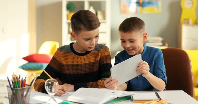 Caucasian little teen boy writing doing homework task while his friend showing him something on the tablet computer in the kid's room.