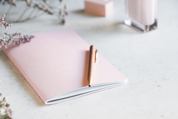 Pink empty paper notebook with golden pen on white table surrounded by flowers and fragrance. The concept of femininity workplace