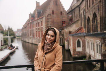 Fototapeta na wymiar Portrait of beautiful woman in brown coat standing on the bridge in Bealgium, Brugge. Female put on a hood