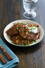 Potato pancakes draniki served with sour cream, chives and bacon on a white plate. Dark wooden table, blue linen napkin, high resolution