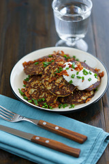 Potato pancakes draniki served with sour cream, chives and bacon on a white plate. Dark wooden table, blue linen napkin, high resolution