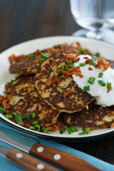 Potato pancakes draniki served with sour cream, chives and bacon on a white plate. Dark wooden table, blue linen napkin, high resolution