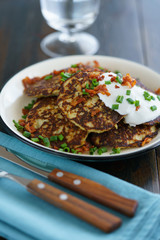 Potato pancakes draniki served with sour cream, chives and bacon on a white plate. Dark wooden table, blue linen napkin, high resolution