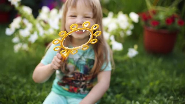 Footage Of Adorable Long Haired Little Girl In Blue Printed T-shirt With Big Beautiful Eyes Blowing Soap Bubbles And Singing Songs To The Camera. She Is Sitting On The Grass In The Backyard Or Garden