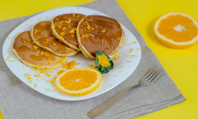 Pancakes with orange for breakfast. A pile of pancakes on a plate with honey and orange on a bright yellow background. View from above. Food flat