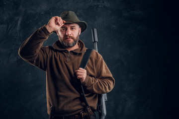 Portrait of a bearded hunter with rifle holding hand on hat and looking at a camera.