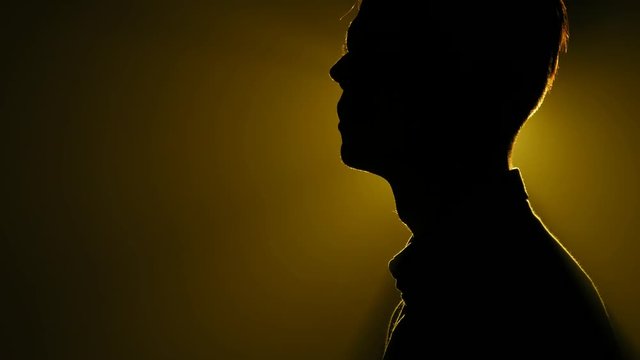 Silhouette of young man standing and looking around in the dark smoky room. Bright yellow lamp behing the man