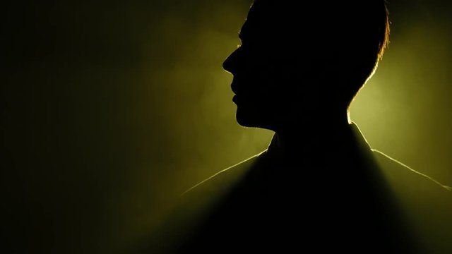 Silhouette of young man standing and looking around in the dark smoky room. Bright yellow lamp behing the man