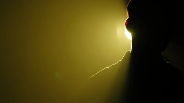Silhouette of young man walking and looking around in the dark smoky room. Bright yellow lamp behing the man