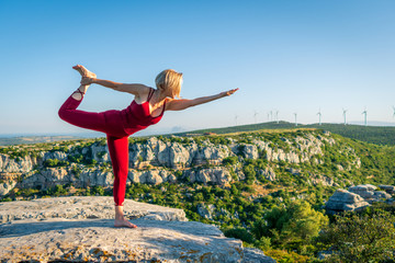 Yoga Fitness Woman Performing Lord of the Dance Pose (Natarajasana) with Bent Foot and Stretched Hand. She is Standing at the Edge of Cliff Overlooking Mountain Range and Ocean in Far Distance.