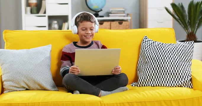 Portrait shot of the joyful Caucasian teen boy in headphones laughing while watching video on the laptop computer and resting on the yellow couch at home.