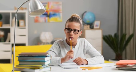 Caucasian pretty small schoolgirl in glasses sitting at the desk in her cozy room and doing homework, thinking with a pencil in hand.