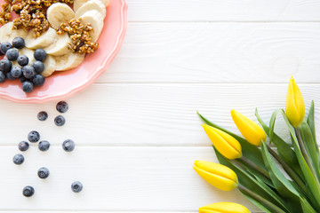 A plate of banana pieces, granola and blueberry on white wooden table. Space for text, copy space. Yellow flowers decorations. Coral living color. 