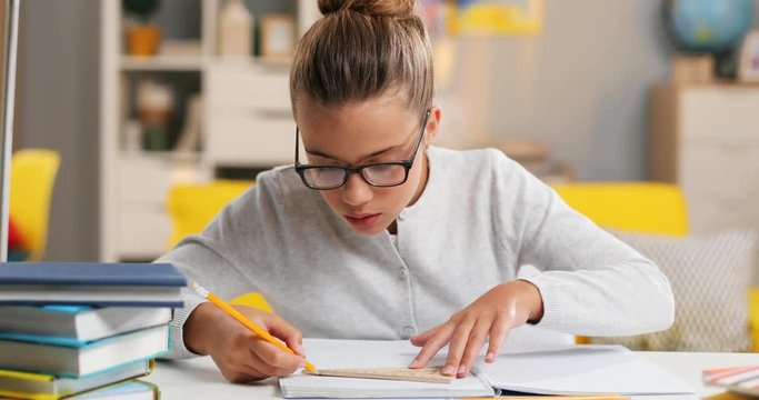 Close up of the Caucasian teenage girl in glasses with a pencil in hand writing down in notebook while doing an exercise of homework in the cozy room.