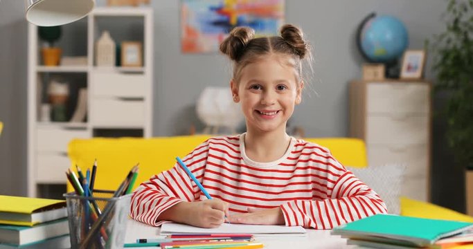 Portrait shot of the Caucasian little and cute girl drawing and coloring a picture in her room and then smiling to the camera.