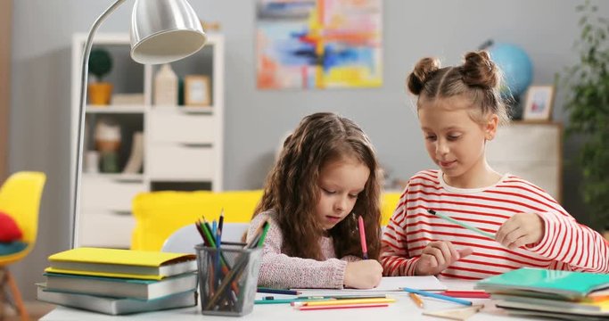 Two cute Caucasian small girls drawing and coloring a picture at the desk in the cozy kid's room at home.