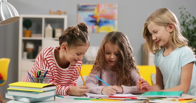 Three cute Caucasian small girls friends drawing and coloring a picture at the desk in the cozy kid's room at home.