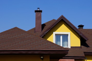 attic of a yellow house with a window and a brown tiled roof against the sky