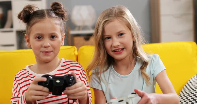 Portrait of the two cheerful and cute Caucasian small sisters sitting on the yellow couch and playing videogames with joysticks in hands in cozy living room.
