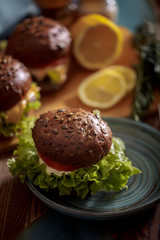 Dark burger with grain bread on dark ceramic plate, salad, rosemary and lemon
