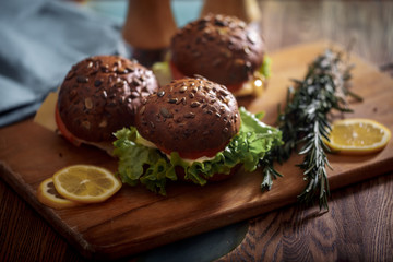 Dark burger with grain bread on dark ceramic plate, salad, rosemary and lemon