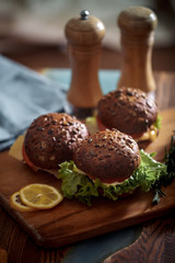 Dark burger with grain bread on dark ceramic plate, salad, rosemary and lemon
